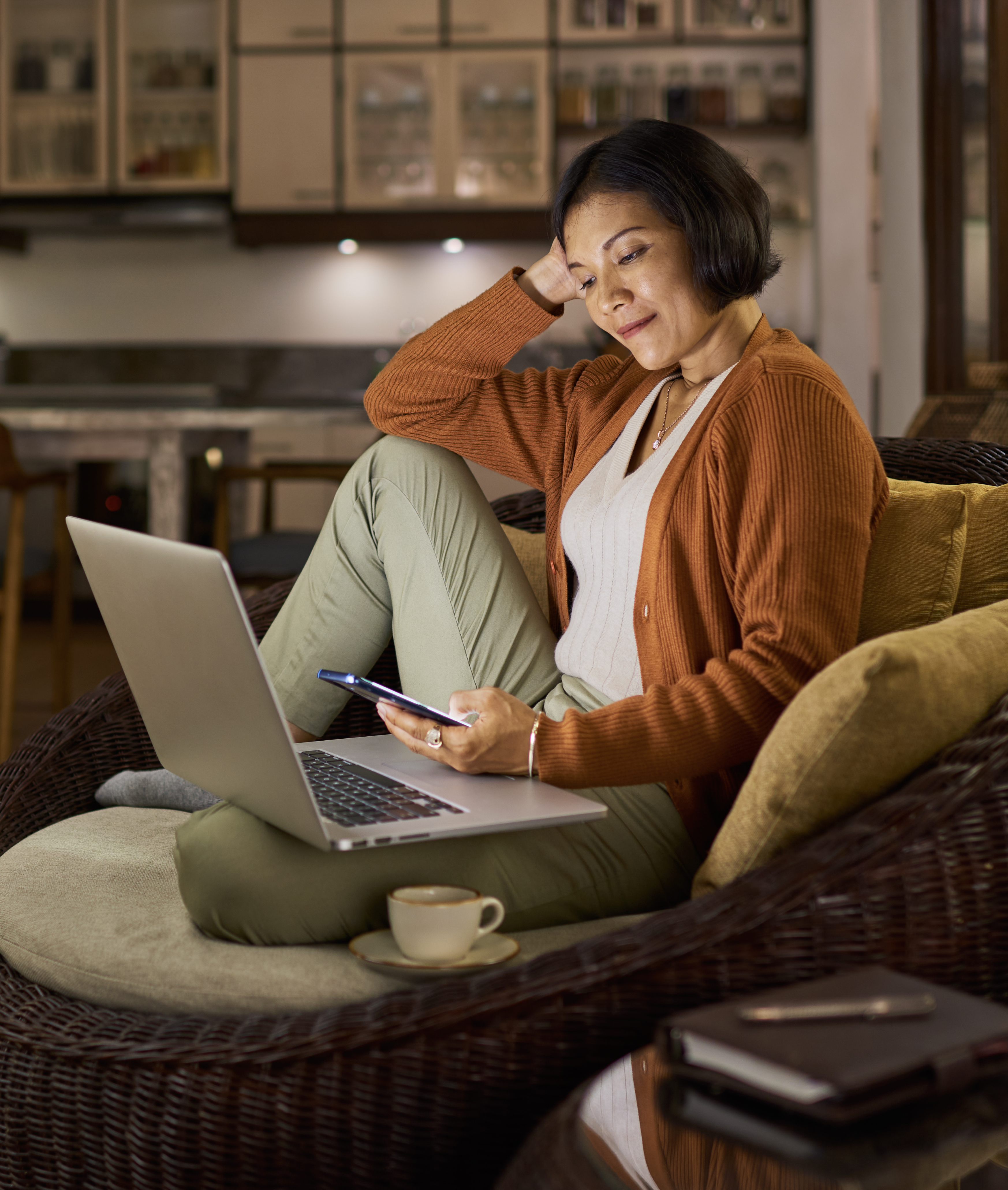 Mature Asian woman working from home sitting in open plan house, using mobile phone and laptop computer at night